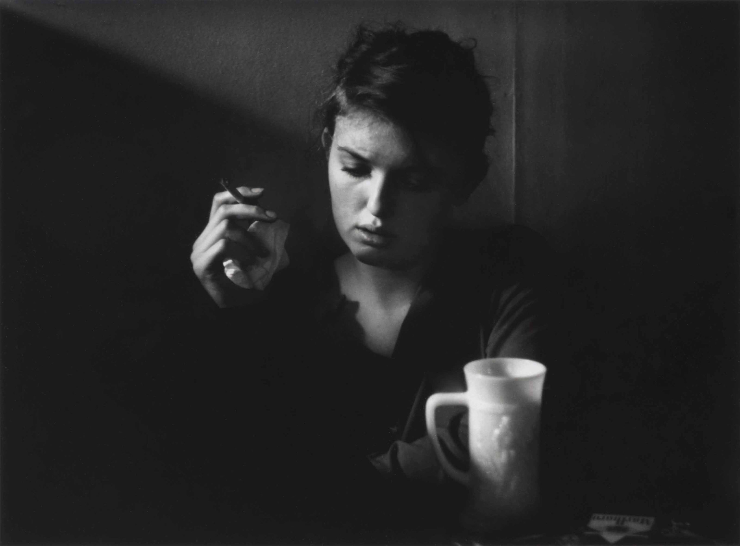 A woman lost in thought over a cup of coffee, cigarette in hand, lit from one side — photograph by Dave Heath