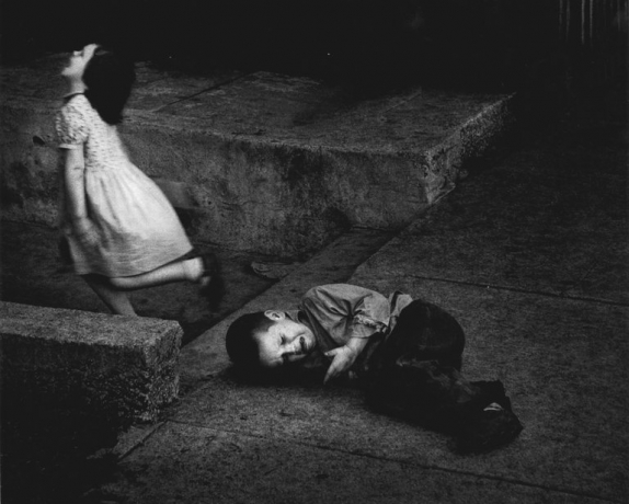 A girl in a white dress runs up stone steps past a boy curled crying on the pavement — photograph by Dave Heath
