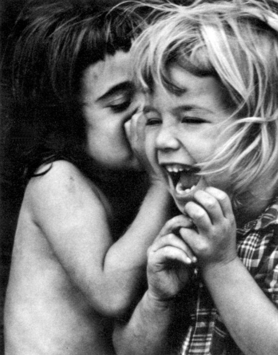 One child whispers into the ear of another, who laughs open-mouthed — photograph by Dave Heath