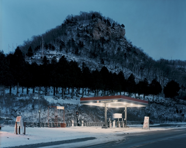 A lit petrol station at dusk beneath a wooded bluff and hillside cemetery — photograph by Alec Soth